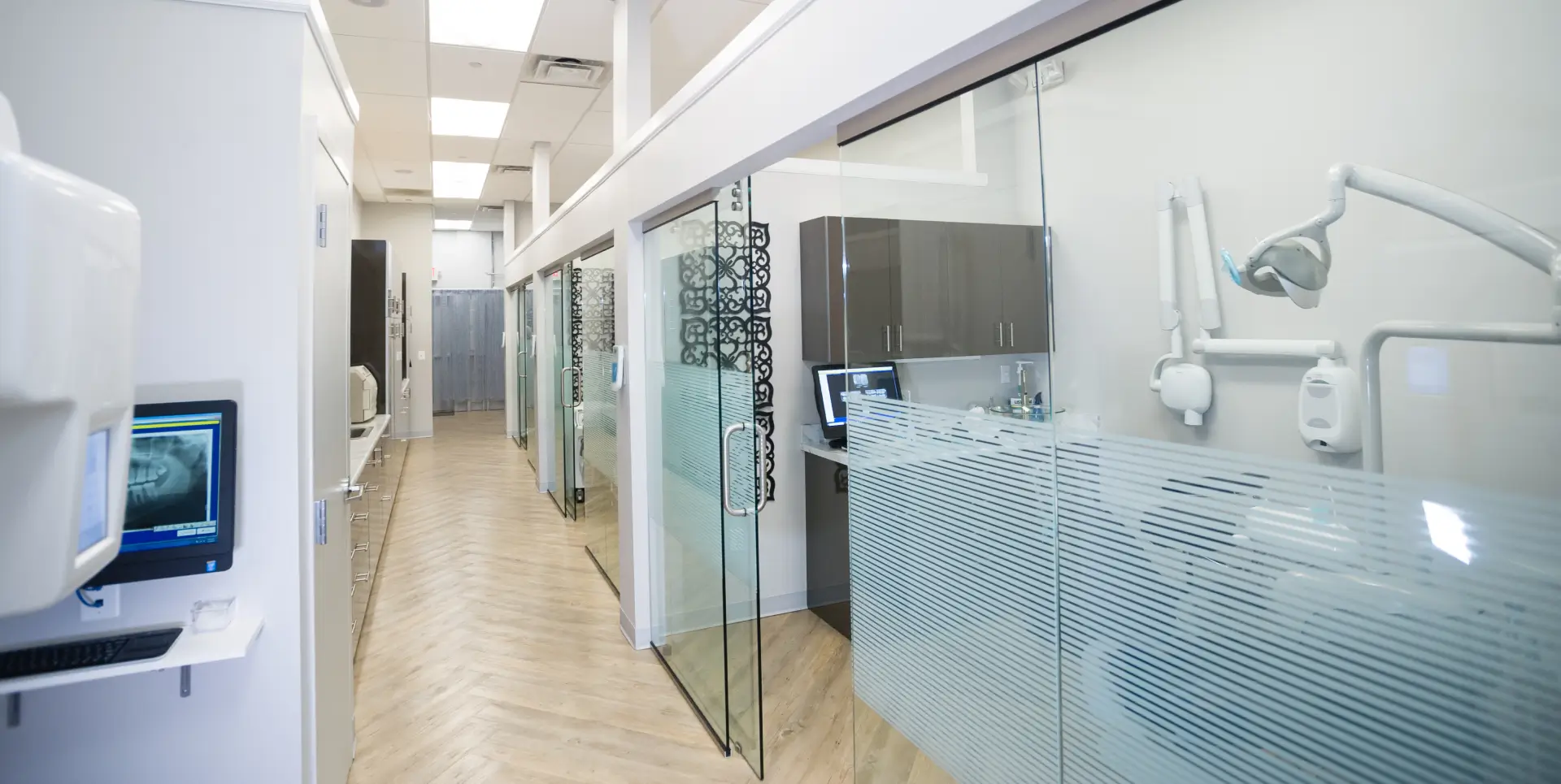 A modern dental office hallway with glass walls, showcasing dental equipment and treatment rooms.