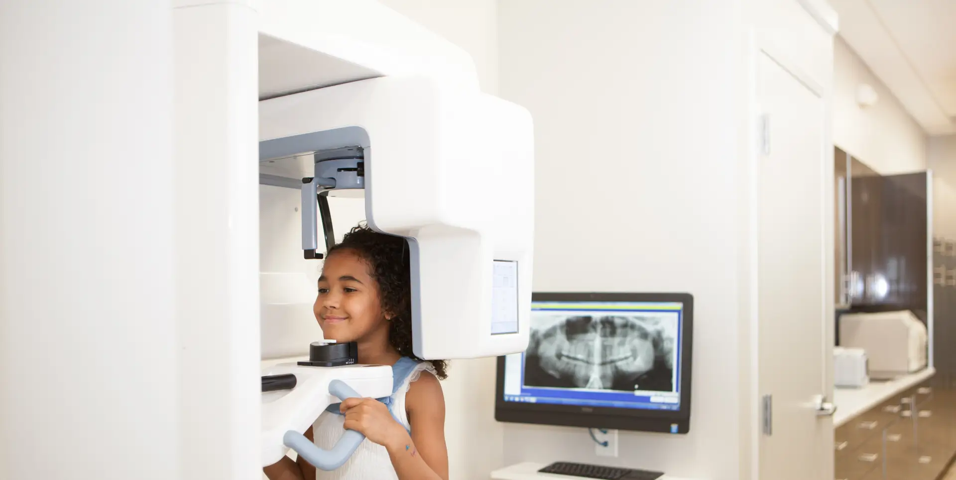 A girl stands inside a dental imaging machine, smiling as an X-ray is taken.