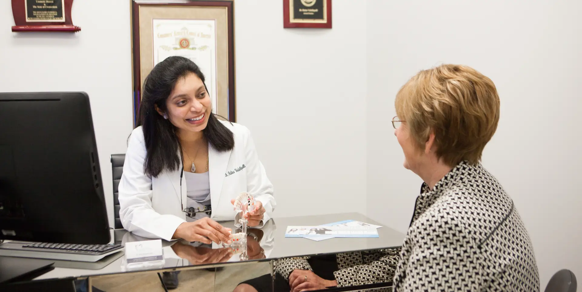 A doctor in a white coat talks with a patient in an office setting.